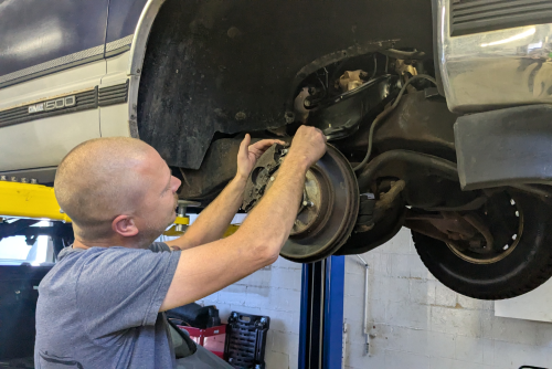 Side profile close up shot of expert specialist technician changes tires, tyres or brake pads of lifted up car, at auto workshop, wears checkered shirt, uniform costume, hat headwear<br />